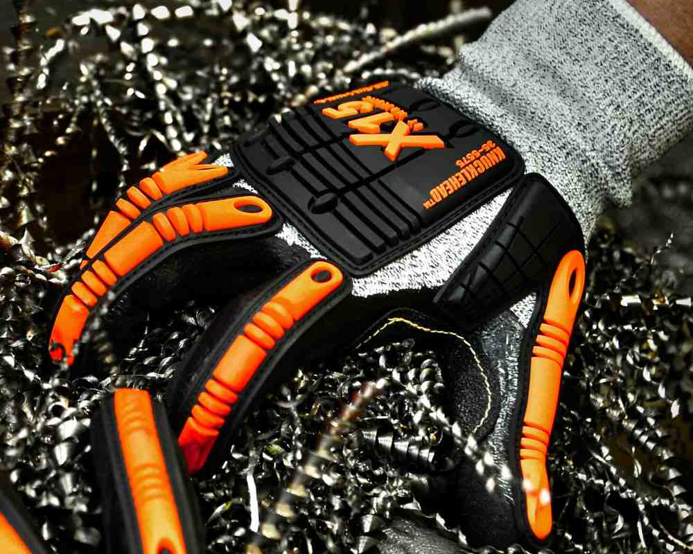 Close-up of a person’s wrist wearing a black and orange grip impact glove, with metal shavings in the background. This setting is a workshop environment.