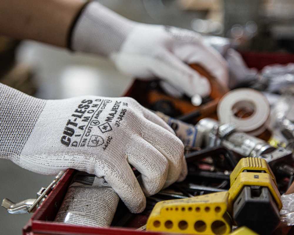 A close-up of a person’s hands wearing white gloves with the word “CUTLESS” printed on them, sorting through various tools and hardware items in a red toolbox. The focus is on the gloved hands, making the background slightly blurred and emphasizing the action of searching for the right tool among screws, nuts, drill bits, and other items.