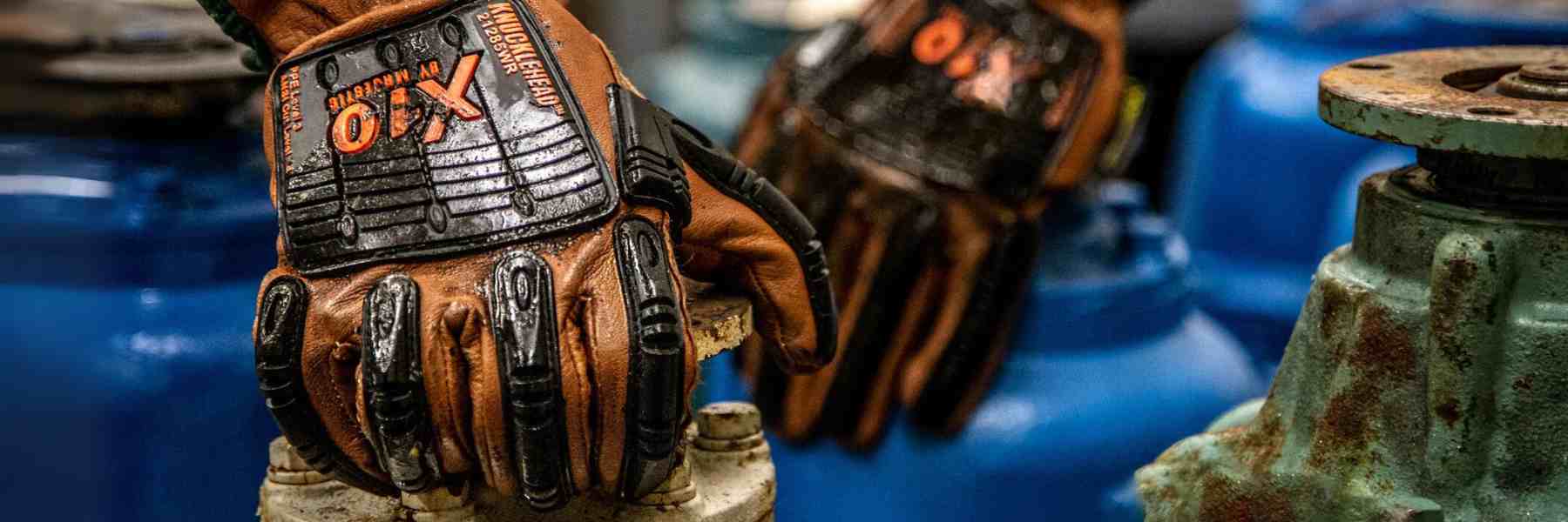 A close-up image of a person’s hands wearing orange and black work gloves with the word “KNUCKLEHEAD” on them. The hands are gripping a metallic, rusted wheel valve attached to what appears to be industrial machinery. The background is blurred but shows hints of blue machinery, suggesting an industrial setting.