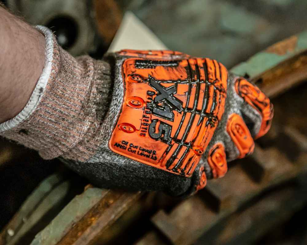 A close-up of a person’s hand wearing a heavy-duty glove with orange and black design elements. The glove has reinforced padding on the back and is labeled “Impact,” designed to protect against mechanical impacts. The person’s wrist is partially visible, showing a gray cuff, likely part of the glove or a sleeve. The background is blurred but appears to be an industrial or workshop setting with metal components, indicating that the glove is used for safety in such environments.