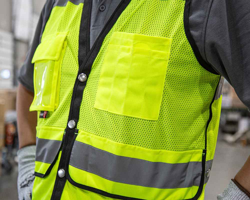 Close-up of a person wearing a high-visibility safety vest with reflective stripes. The vest is fluorescent yellow-green with mesh fabric and has a vertical reflective stripe on the front and horizontal stripes on the shoulder areas. The person is also wearing gray gloves, and the background is an industrial setting.